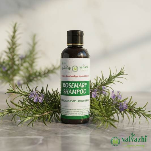 Nalvazhi Rosemary Shampoo dark bottle surrounded by fresh rosemary sprigs with purple-blue flowers on light grey marble with soft natural lighting